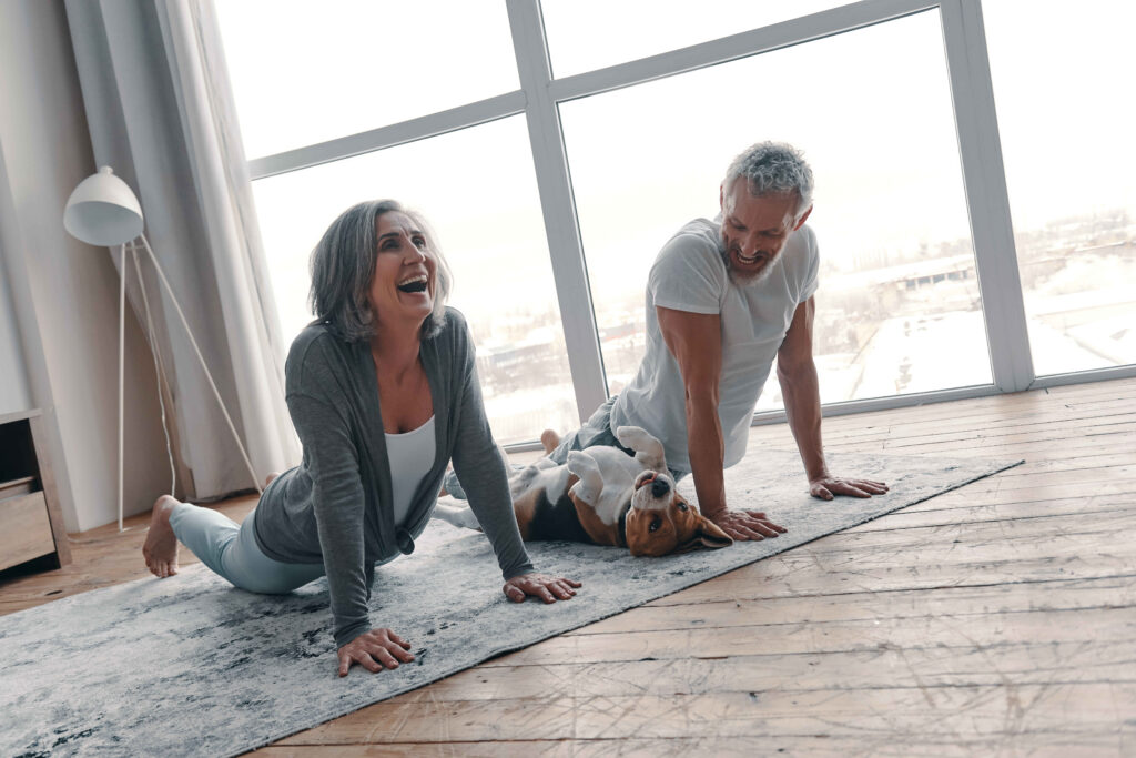 Active senior couple in sports clothing doing yoga and smiling while spending time at home with their dog